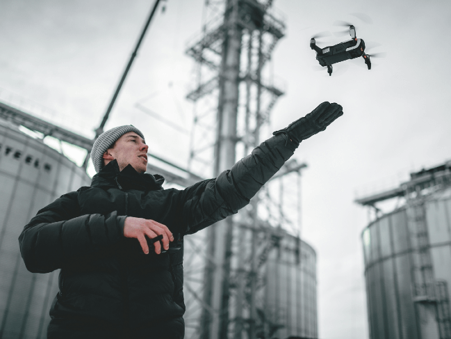 A person dressed in a black winter jacket and grey beanie is flying a small drone, which hovers above their gloved hand, near industrial structures and silos. The overcast sky adds a dramatic backdrop as they stand outdoors, embodying the essence of what defines the modern use of drones.